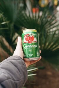 Person holding a Guaraná Antarctica soda can outdoors with a green background.